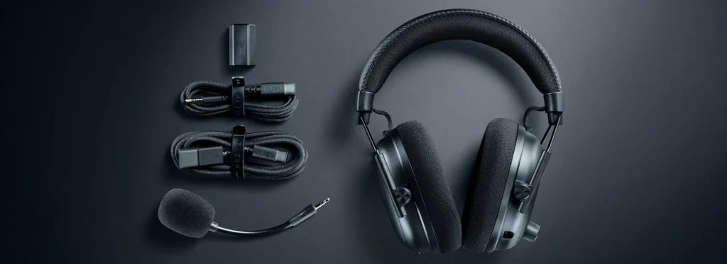 The Razer Blackshark V3 Pro wireless headset laid out on a dark surface with all of its included accessories: the detachable microphone, wireless dongle, and charging cables.