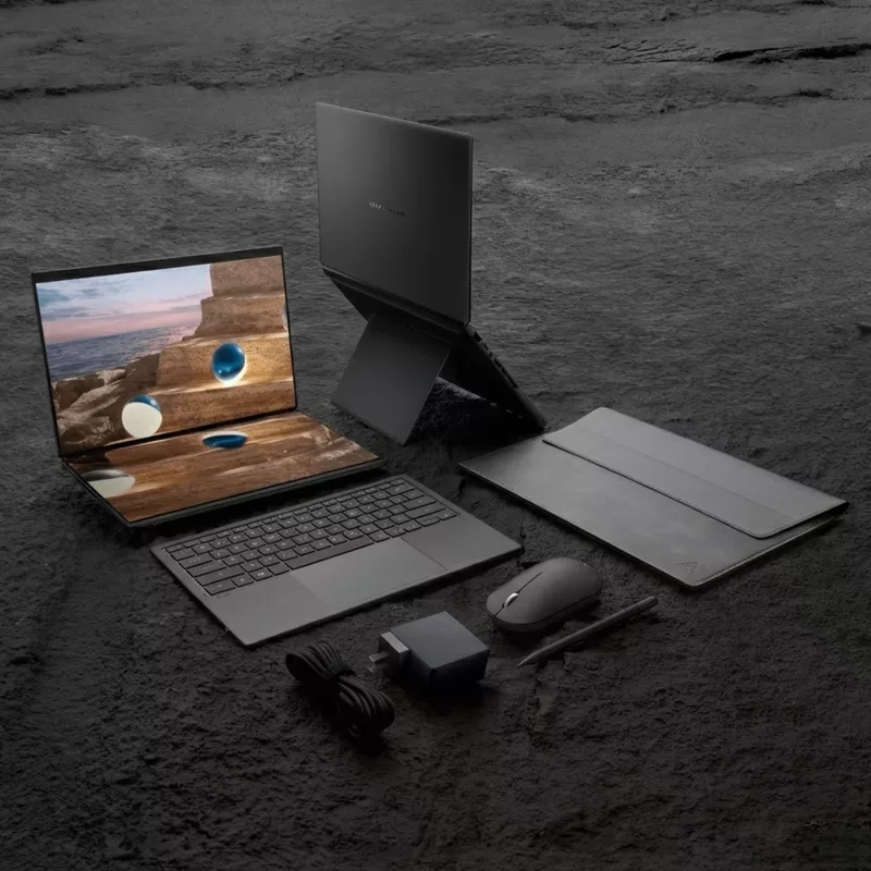 The full ASUS Zenbook Duo package displayed on a stone surface, including the laptop, sleeve, stylus pen, charger, and mouse.